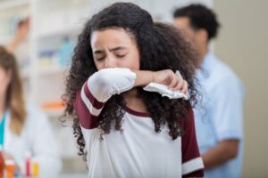 Pediatric patient coughs into crook of elbow holding tissue in medical office